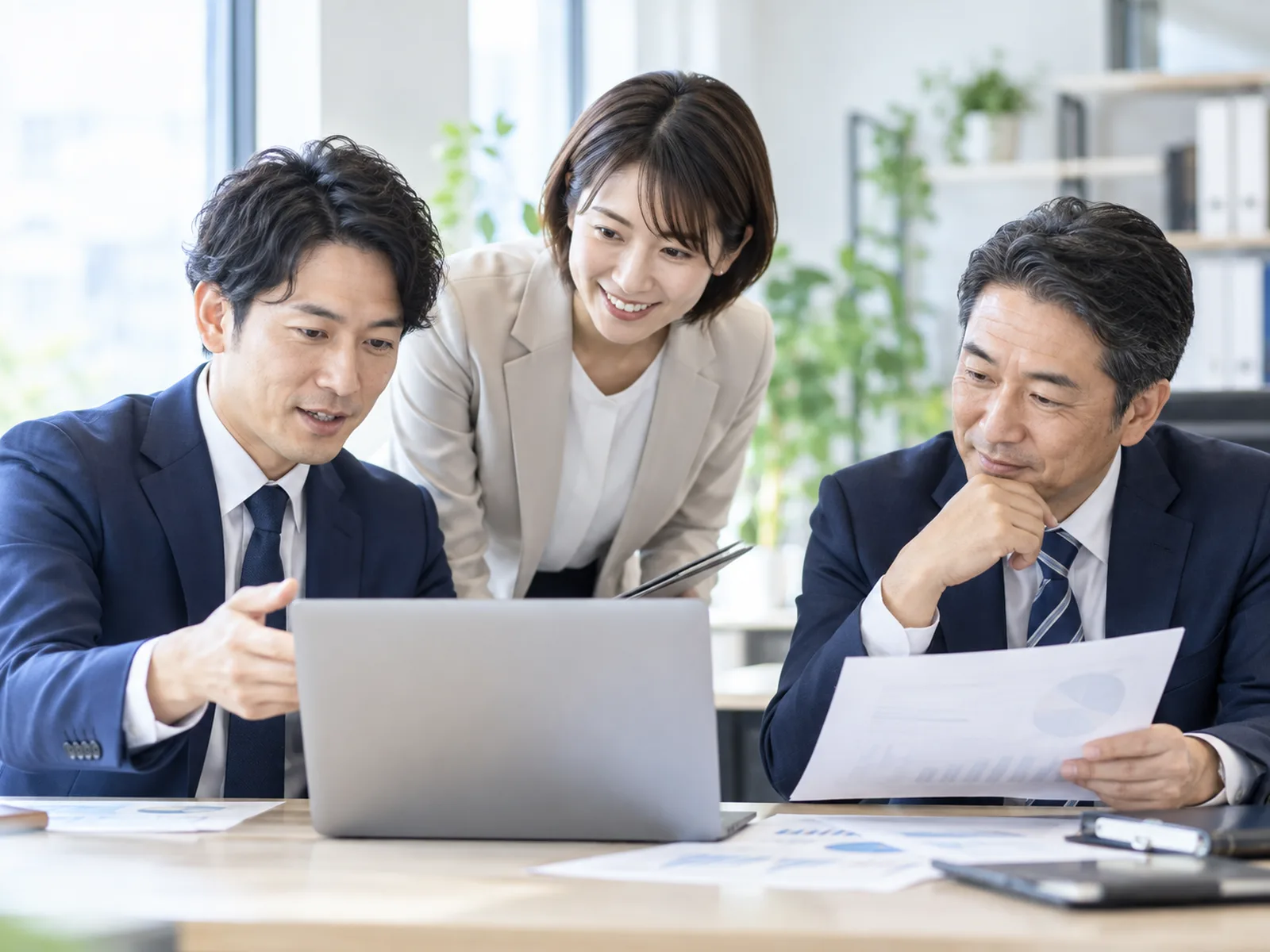 明るい事務所でPCと資料を囲み、協力して働く3人
